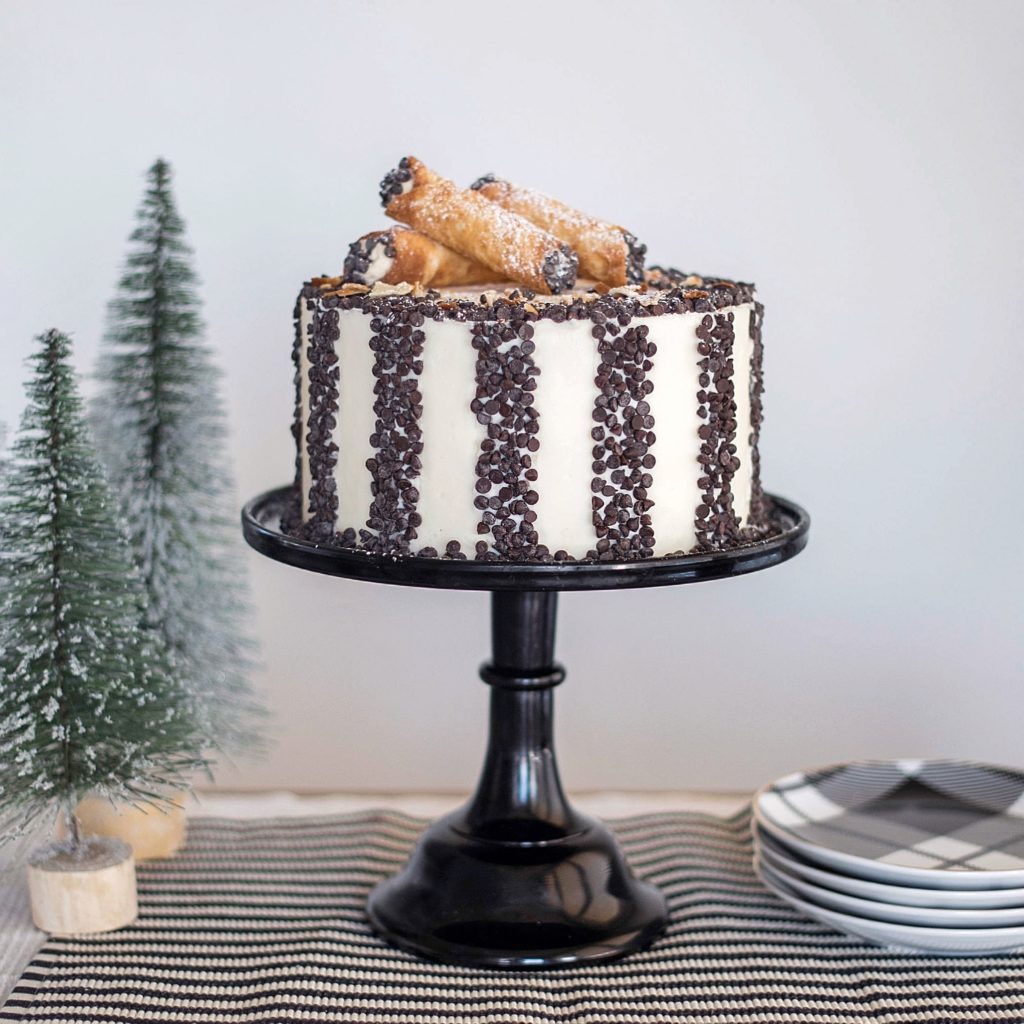 Delicious white cake layers with a hint of cinnamon, a creamy sweet ricotta filling with chocolate chips and vanilla buttercream. #cannoicake #cannolirecipe #homemadecannoli #christmascake #christmasdessert #holidaydessert #christmasdessertideas #christmascakeideas #cakebycourtney
