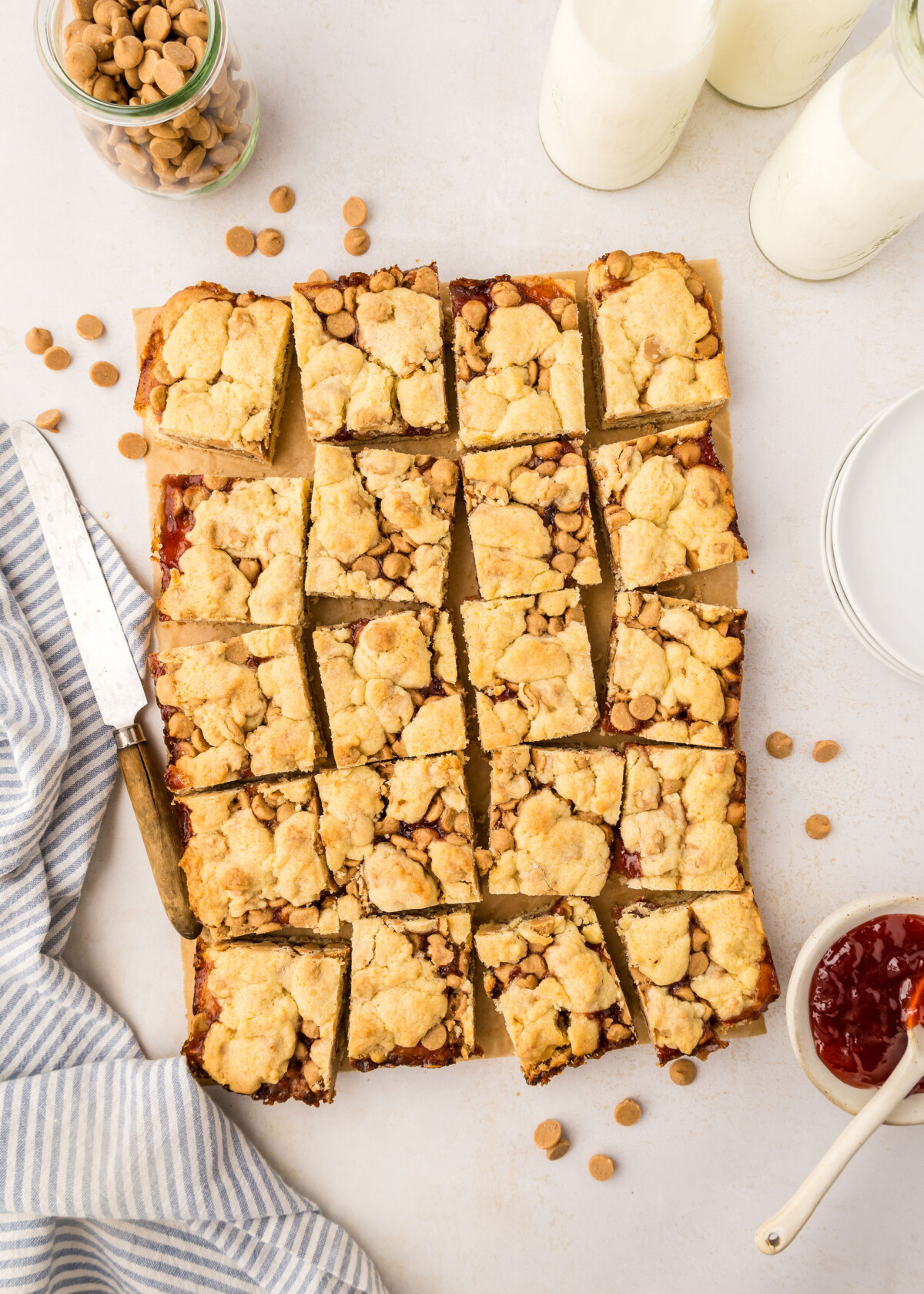 Peanut butter and jelly bars cut into squares on a table with milk in cups.