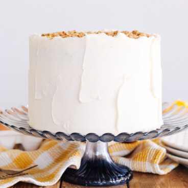 Peach Biscoff Cake on a blue glass cake stand with a kitchen towel and plates on the table.