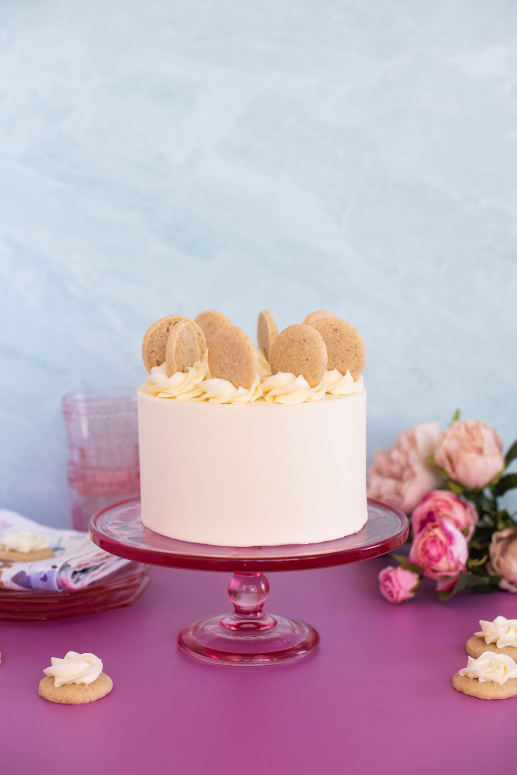Small two-layer cake on a pink cake stand with cookies on the table.