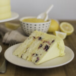 A slice of lemon blueberry cake on a plate with lemon curd and lemons nearby.