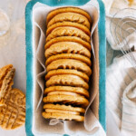 A stack of peanut butter sandwich cookies in a bread dish with a towel and whisk next to it.