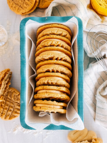 A stack of peanut butter sandwich cookies in a bread dish with a towel and whisk next to it.