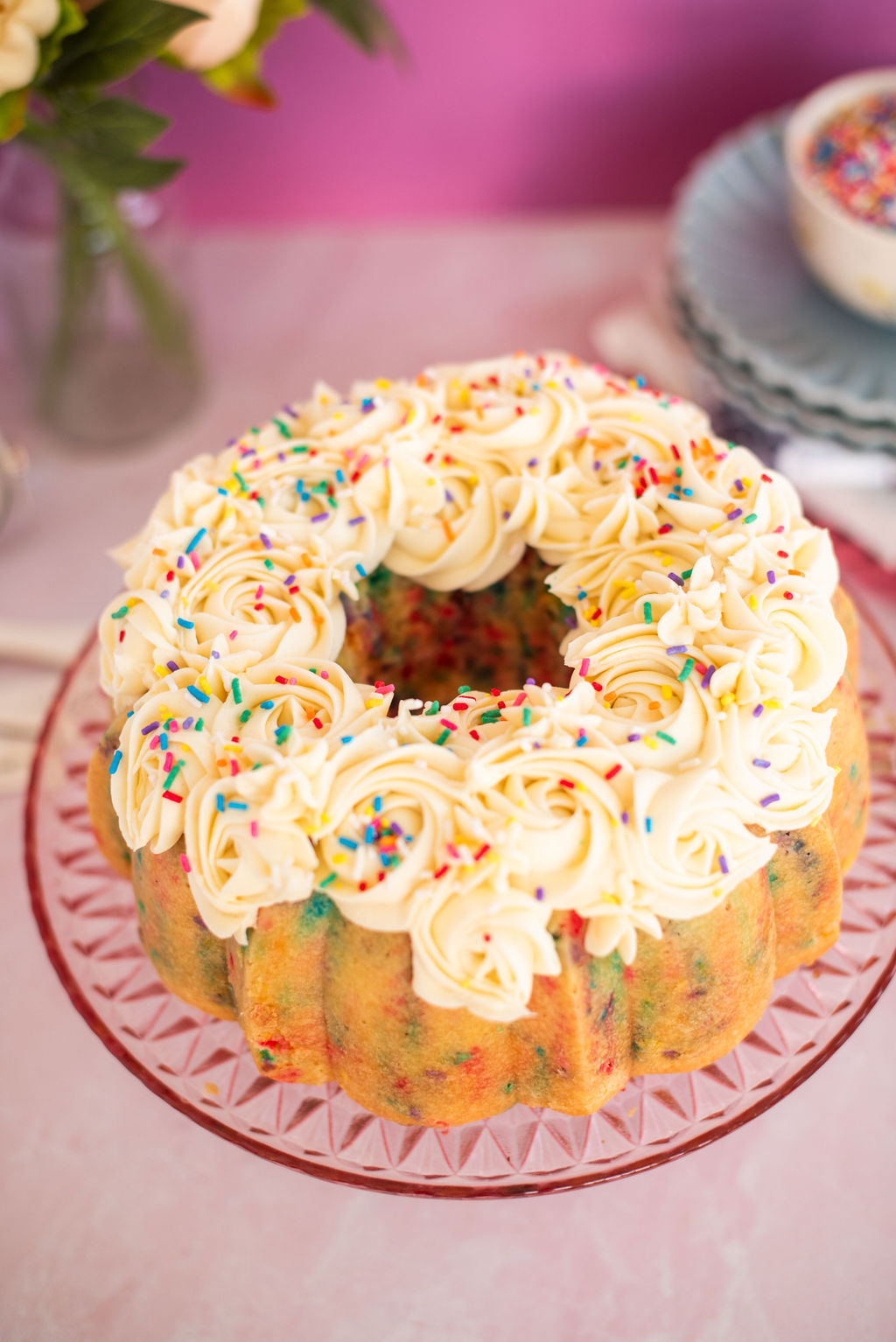 A confetti bundt cake with vanilla buttercream on a pink cake stand.