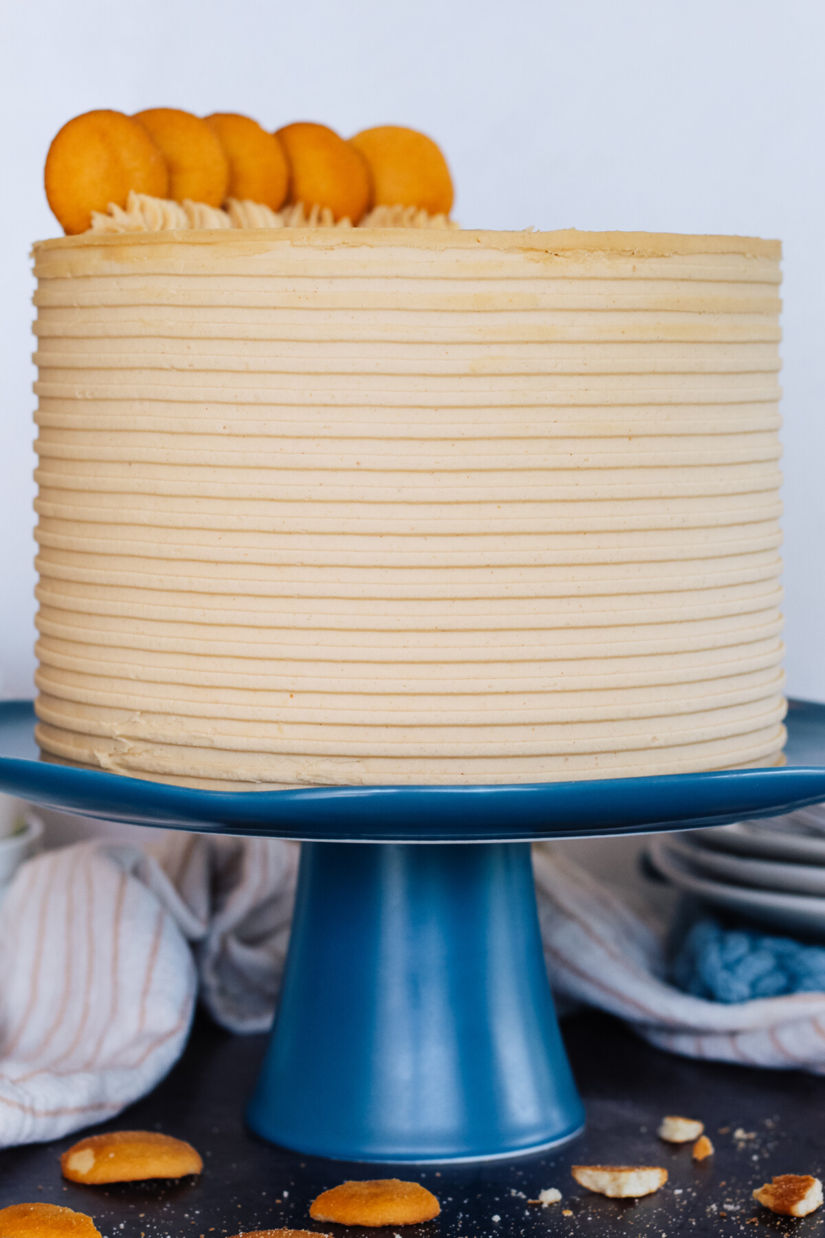 A tall layered cake on a blue cake stand with cookies and plates next to it.