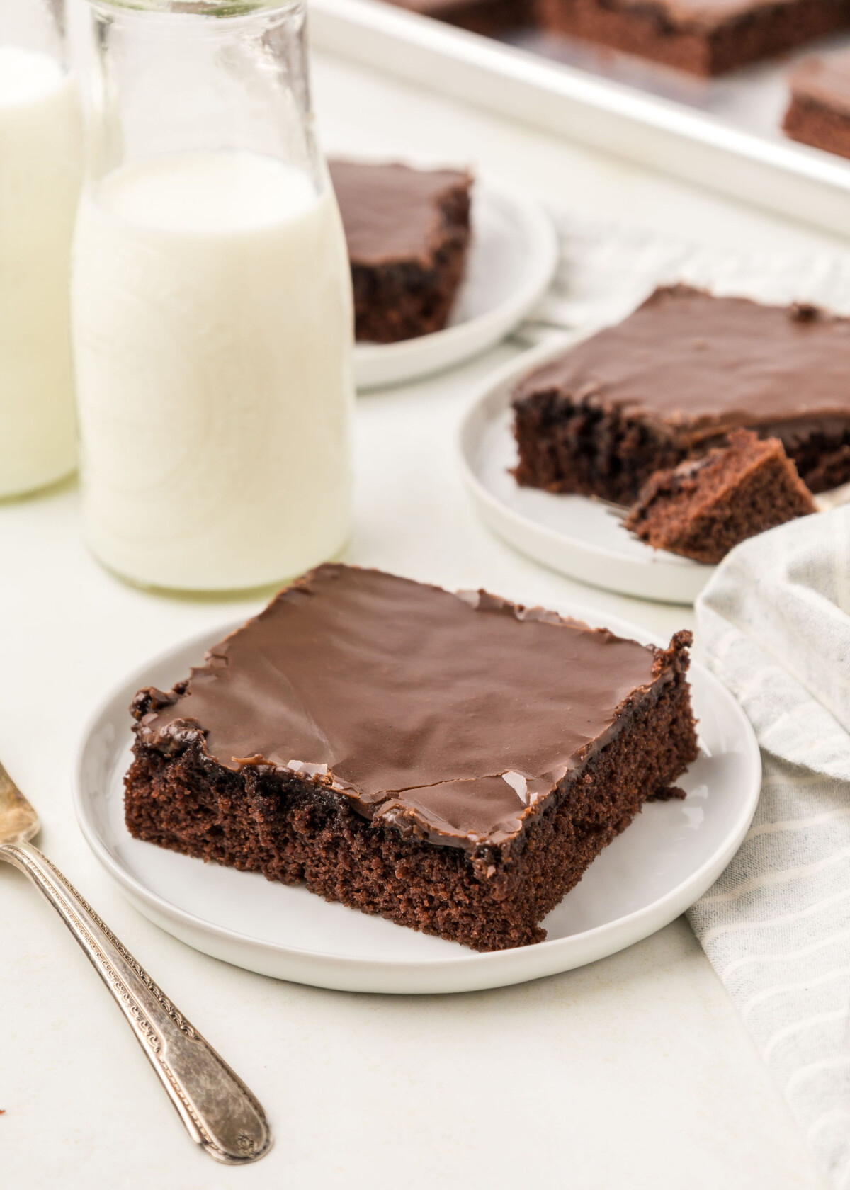 Three slices of Texas Sheet Cake on plates with milk in bottles.