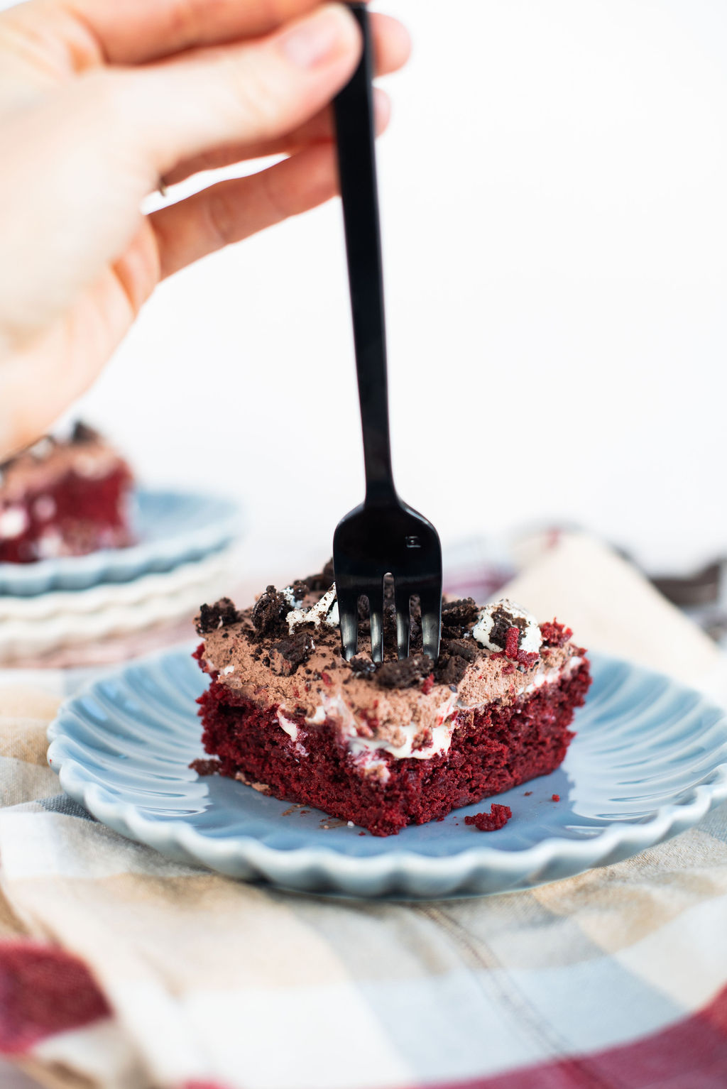 A slice of red velvet Oreo poke cake on a blue scalloped plate with a person taking a bite out of it with a black fork.